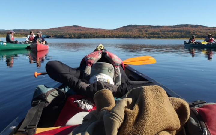From the back of a canoe, you can see the person in front leaning back in relaxing fashion. There are additional canoes floating on calm water in the background.